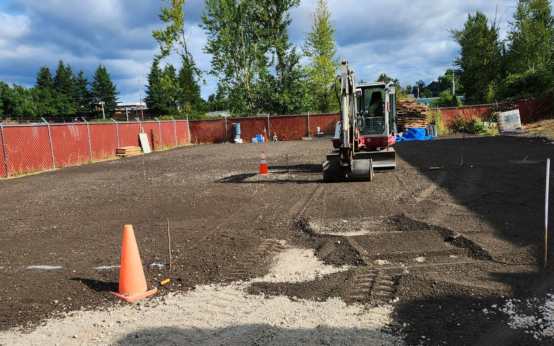 An excavator on a graded dirt lot; orange traffic cone in foreground; a fence and trees in the background.