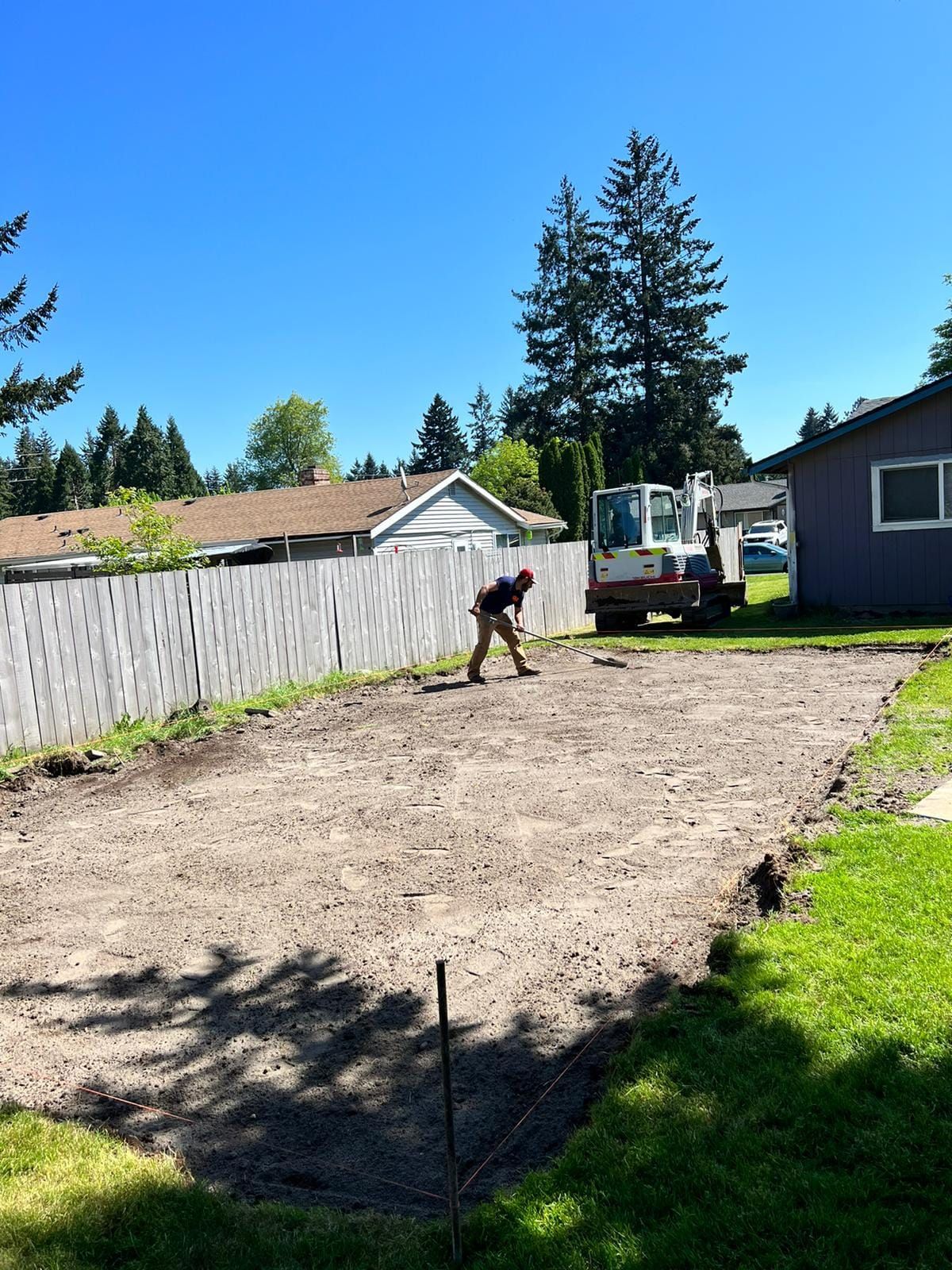 A person raking dirt in a cleared area. Construction scene with a backhoe, houses, and fence under blue sky.