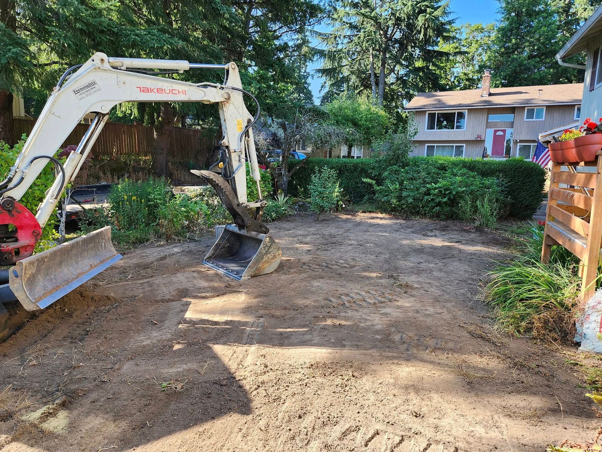 Mini excavator in a yard, soil tilled. Buildings and bushes in background.
