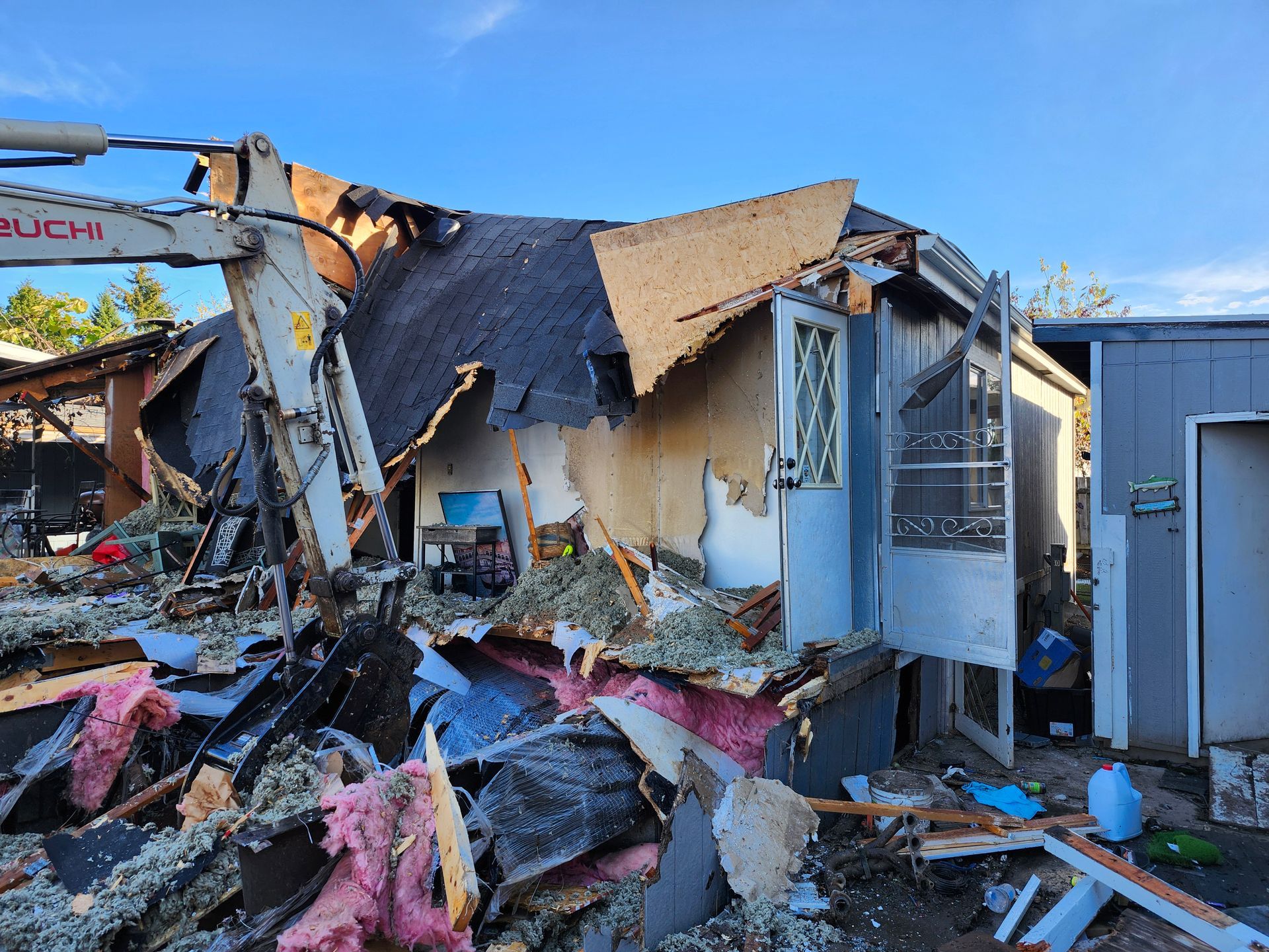 Demolition of a small building, excavator arm visible, debris scattered, blue sky.