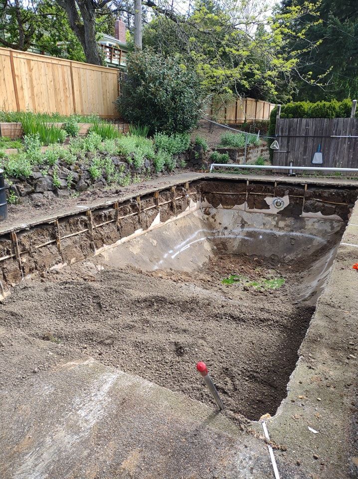 Construction site of an empty pool. Brown dirt and gray concrete. Fenced backyard.