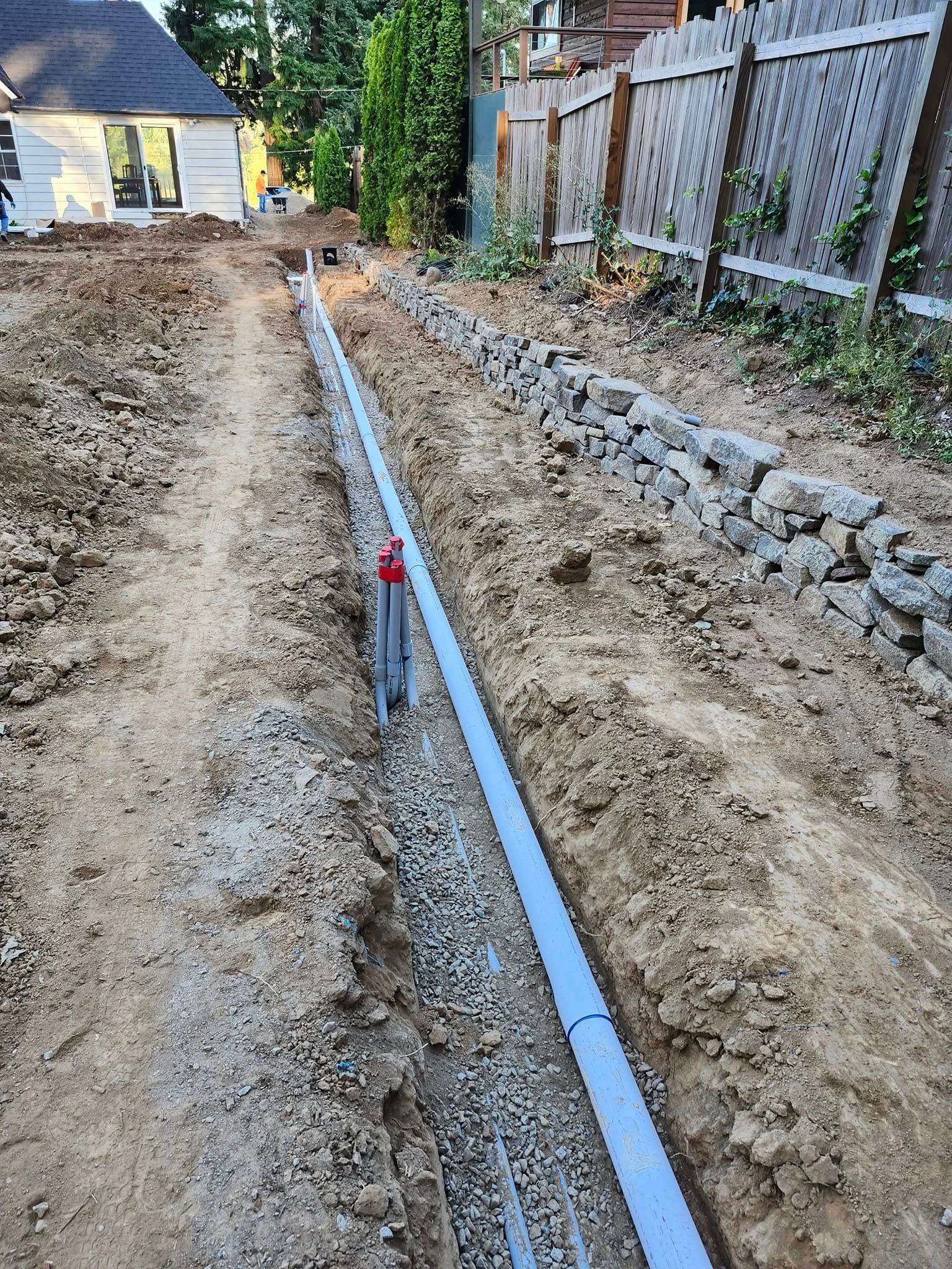 Trench dug for a drain pipe along a dirt pathway, with a retaining wall of gray stones on the right.