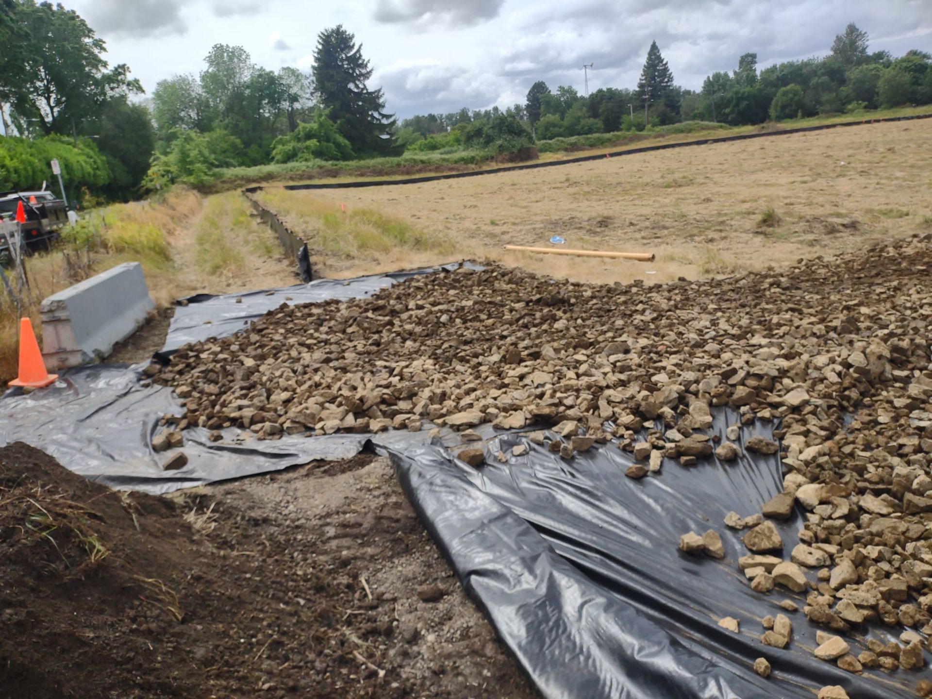 Construction site with black tarp covered in rocks, near a field and a road.
