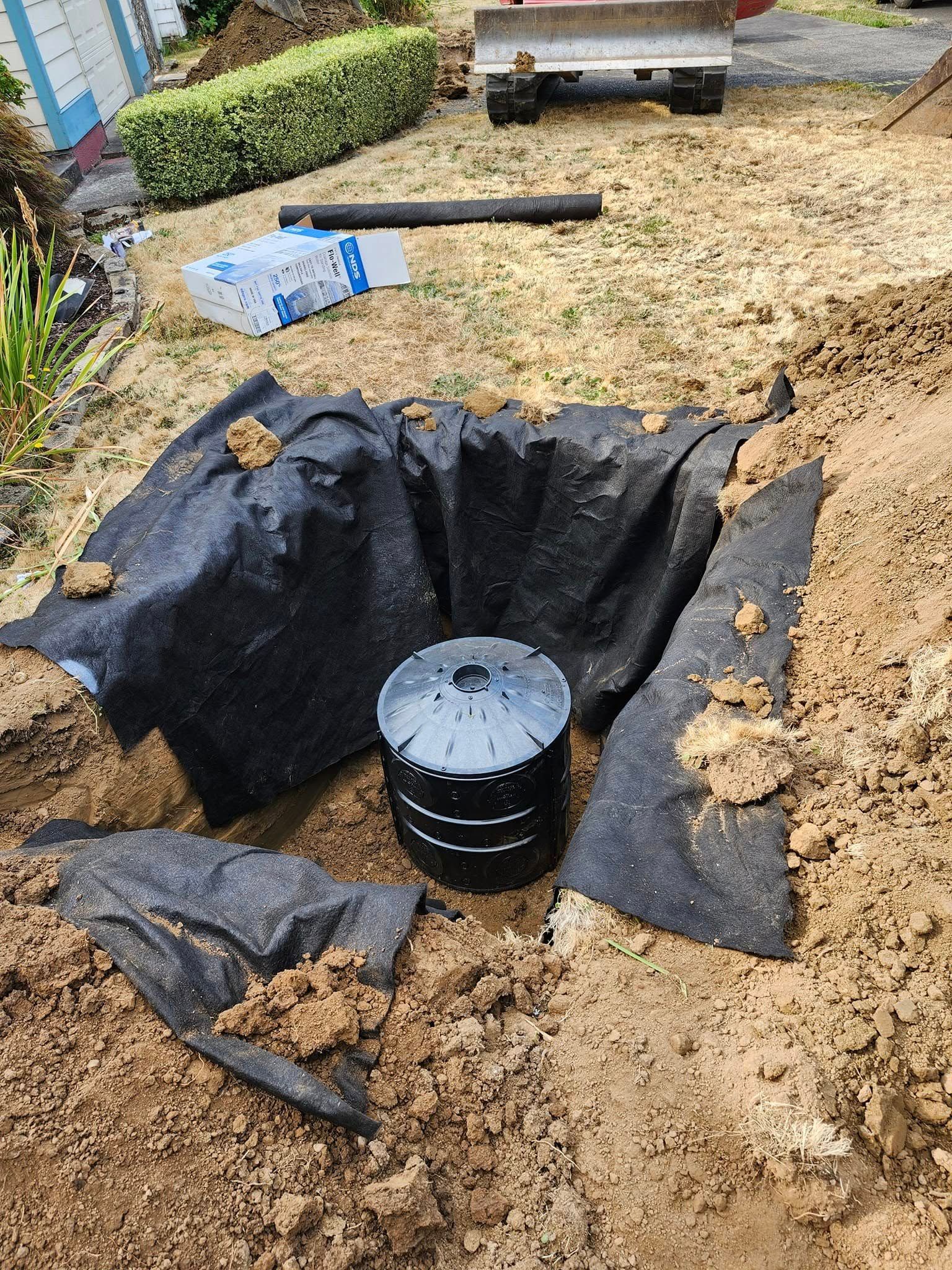 A dark, cylindrical structure is in a dug-out hole, lined with black fabric, outdoors.