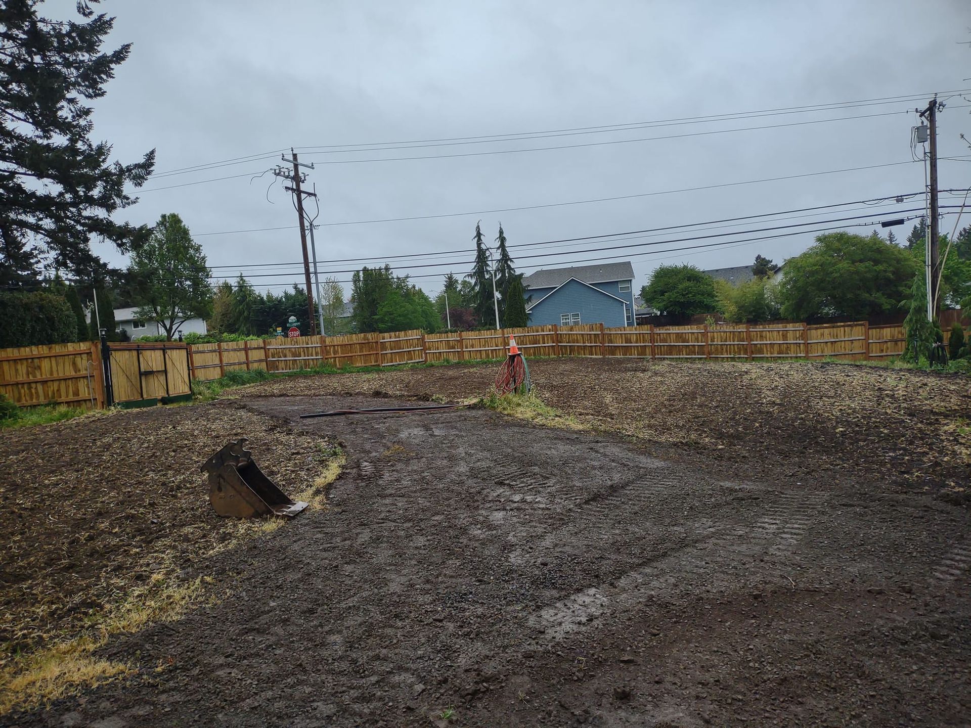 A muddy lot enclosed by a wooden fence under a cloudy sky. Power lines, trees, and houses are visible.
