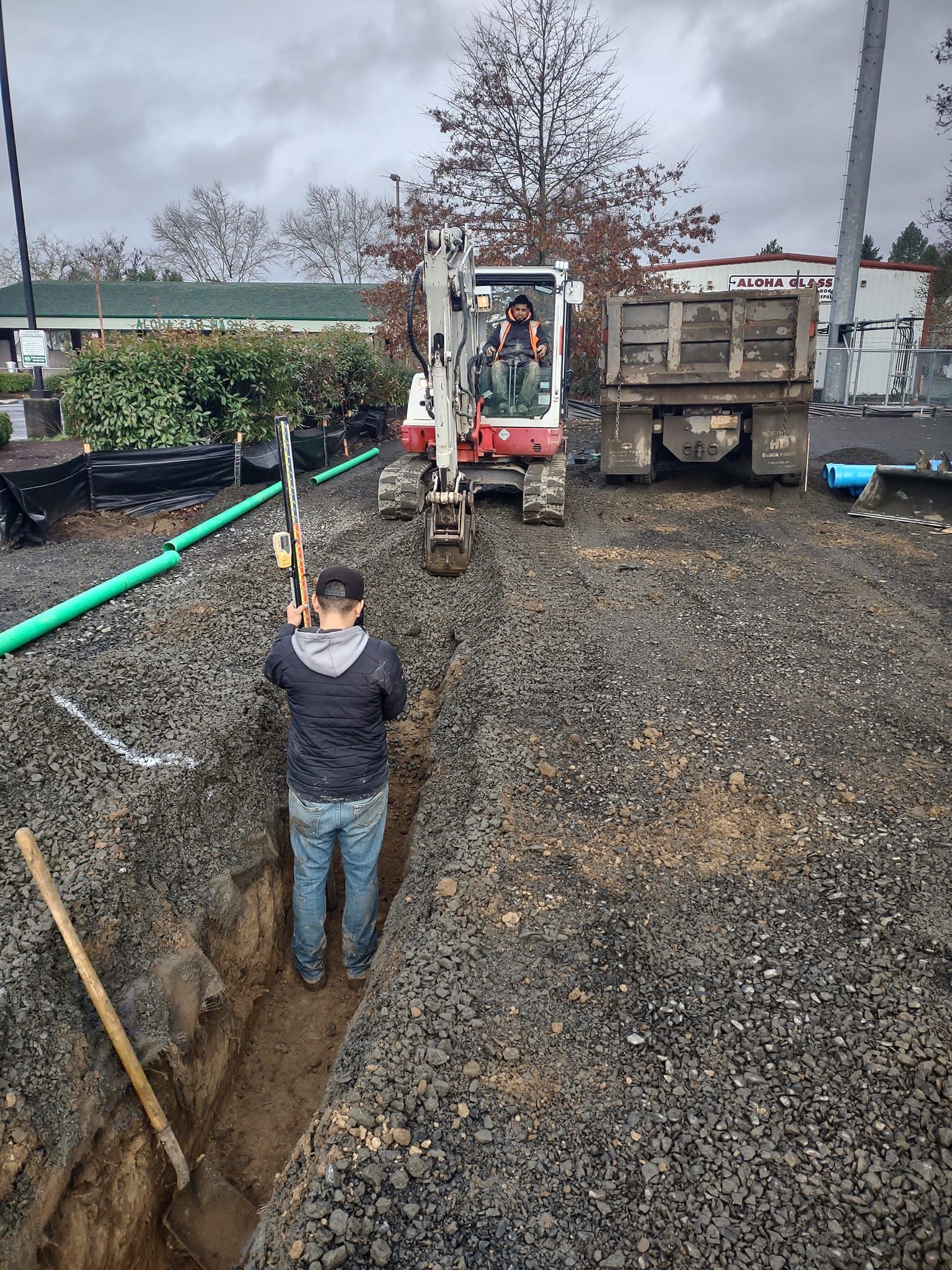 Man digging trench with tools as excavator and truck await on construction site.