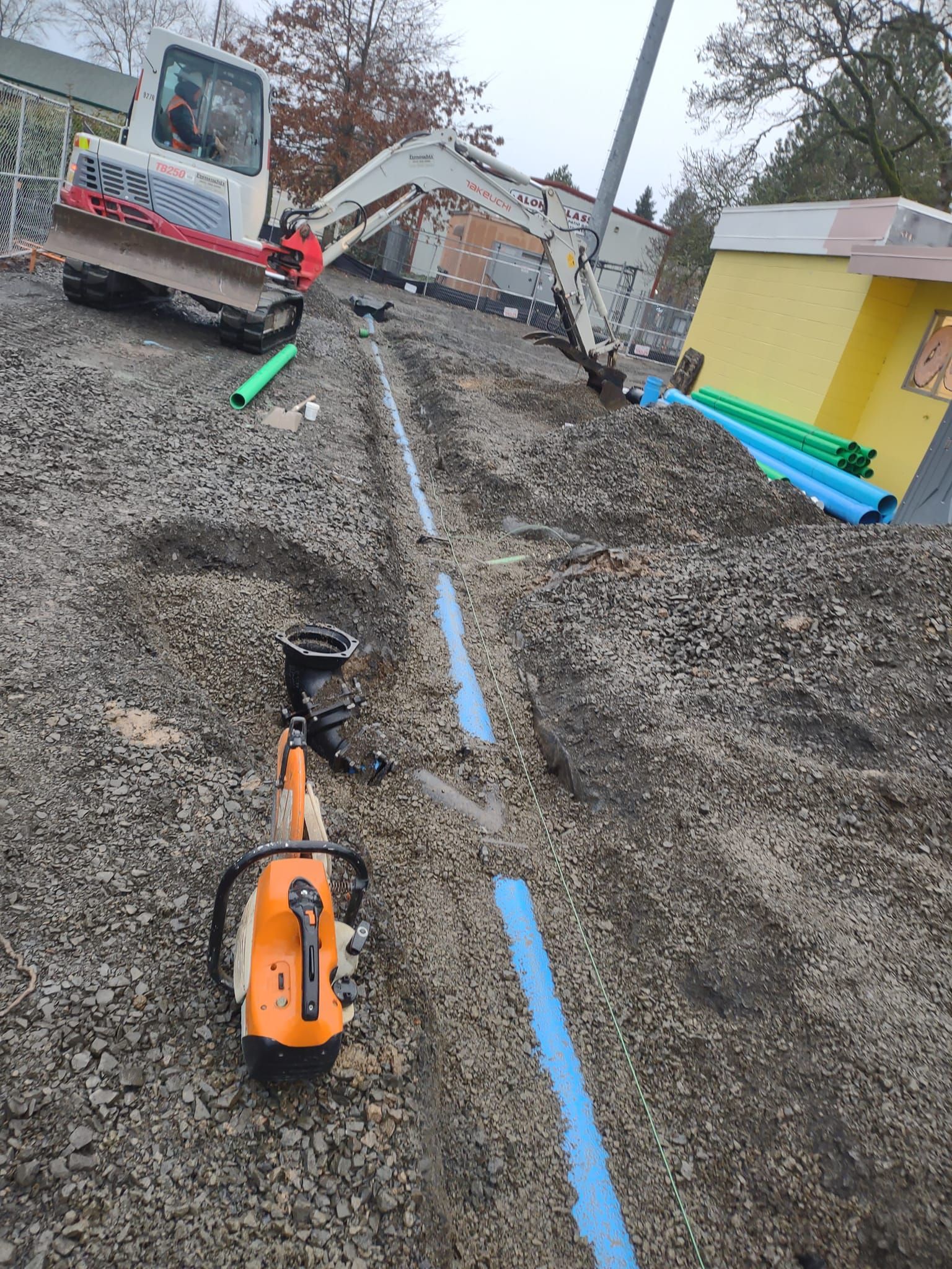 Trench dug with blue pipes, orange chainsaw, small excavator, and building under construction.
