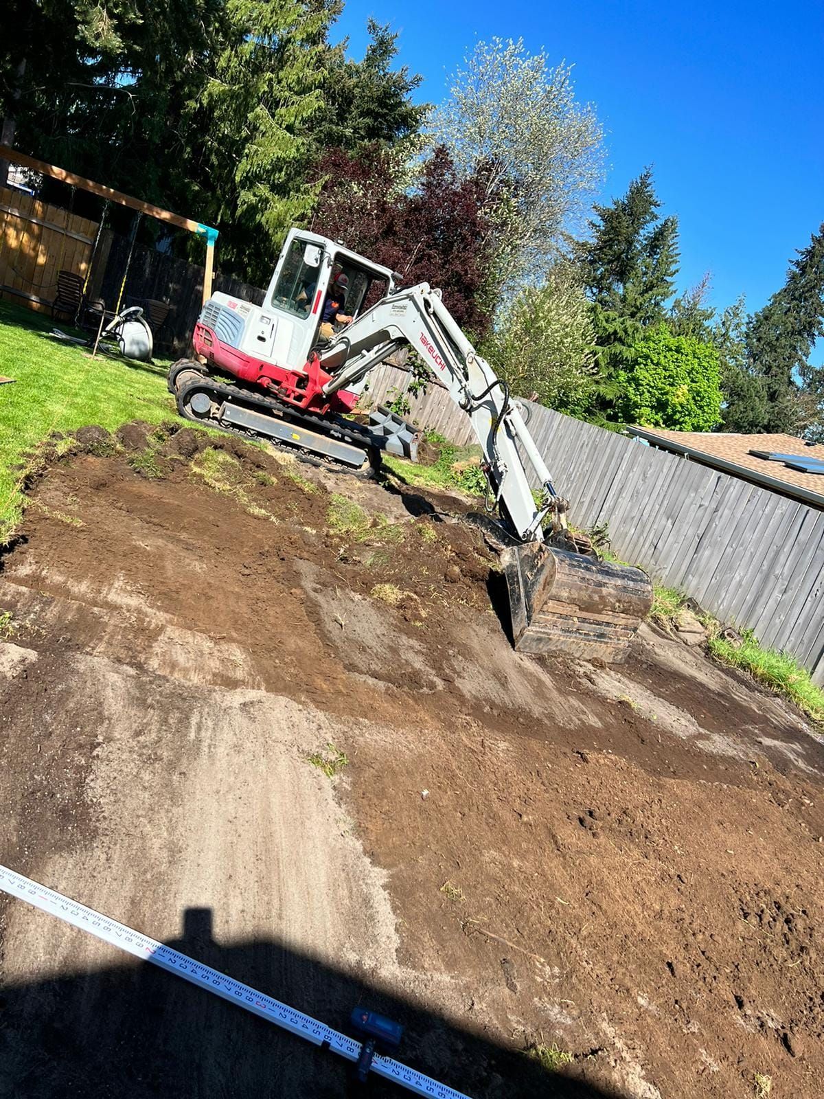 Excavator on a dirt hill next to a fence. Machine is white and red; sky is blue.