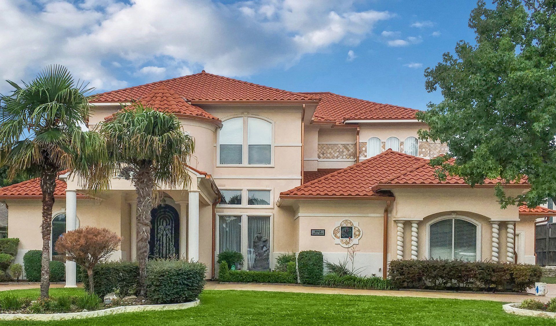 Two-story beige house with a red tile roof, arched windows, and palm trees on a green lawn under a blue sky.