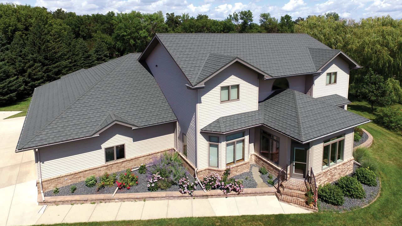 Two-story beige house with gray roof and stone accents, landscaped with flowers, trees in the background.