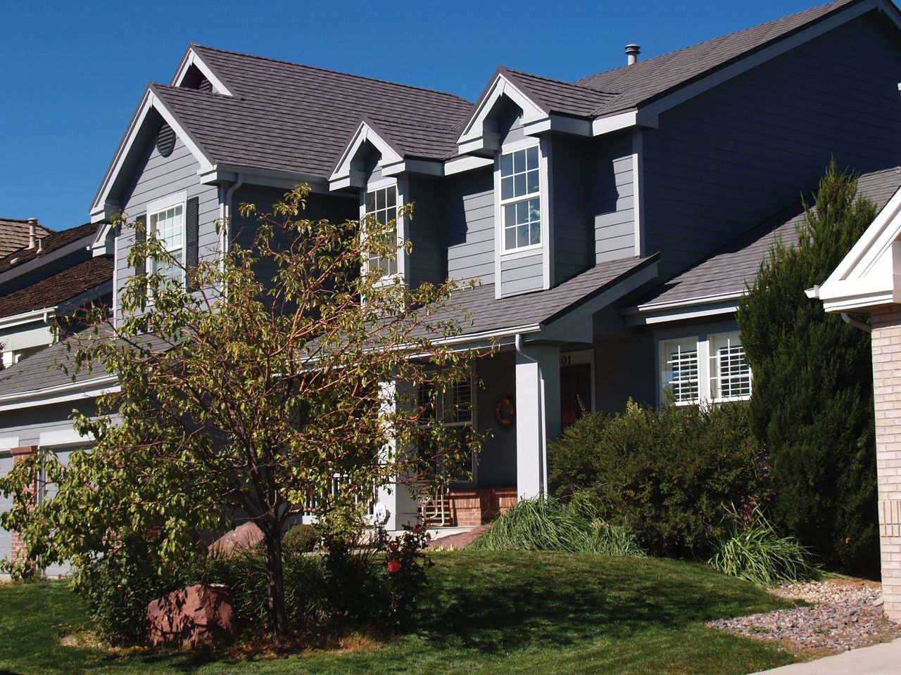Gray two-story house with multiple dormers and dark gray roof, surrounded by green grass, bushes, and a small tree under a bright blue sky.