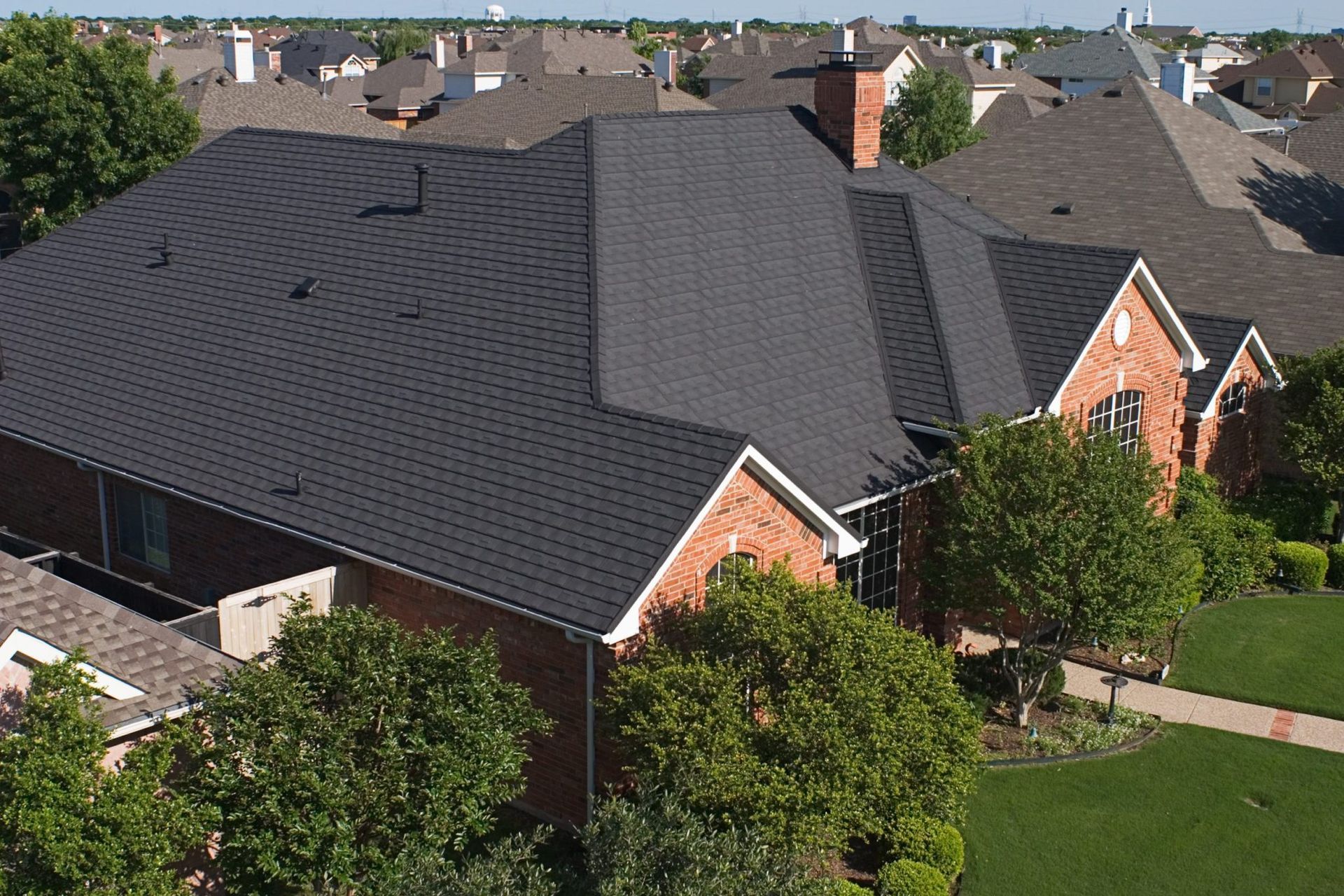 A brick house with a black roof sits in a neighborhood. Green trees surround the house.