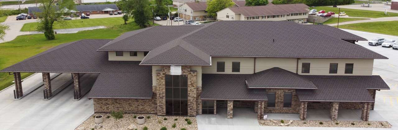 A large, modern building with a dark shingled roof, stone accents, and a spacious parking lot in a grassy area.