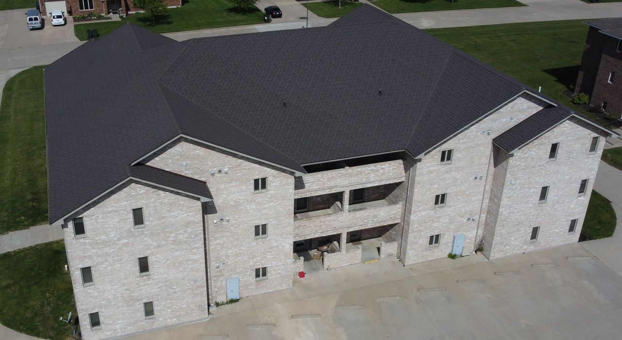 Two-story brick apartment building with a dark gray roof, viewed from above, surrounded by grass.