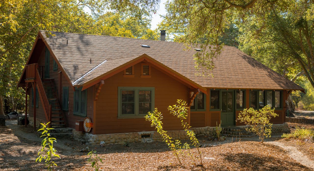 Brown cottage with a striped roof, surrounded by trees and foliage. The house has a porch and a small chimney.