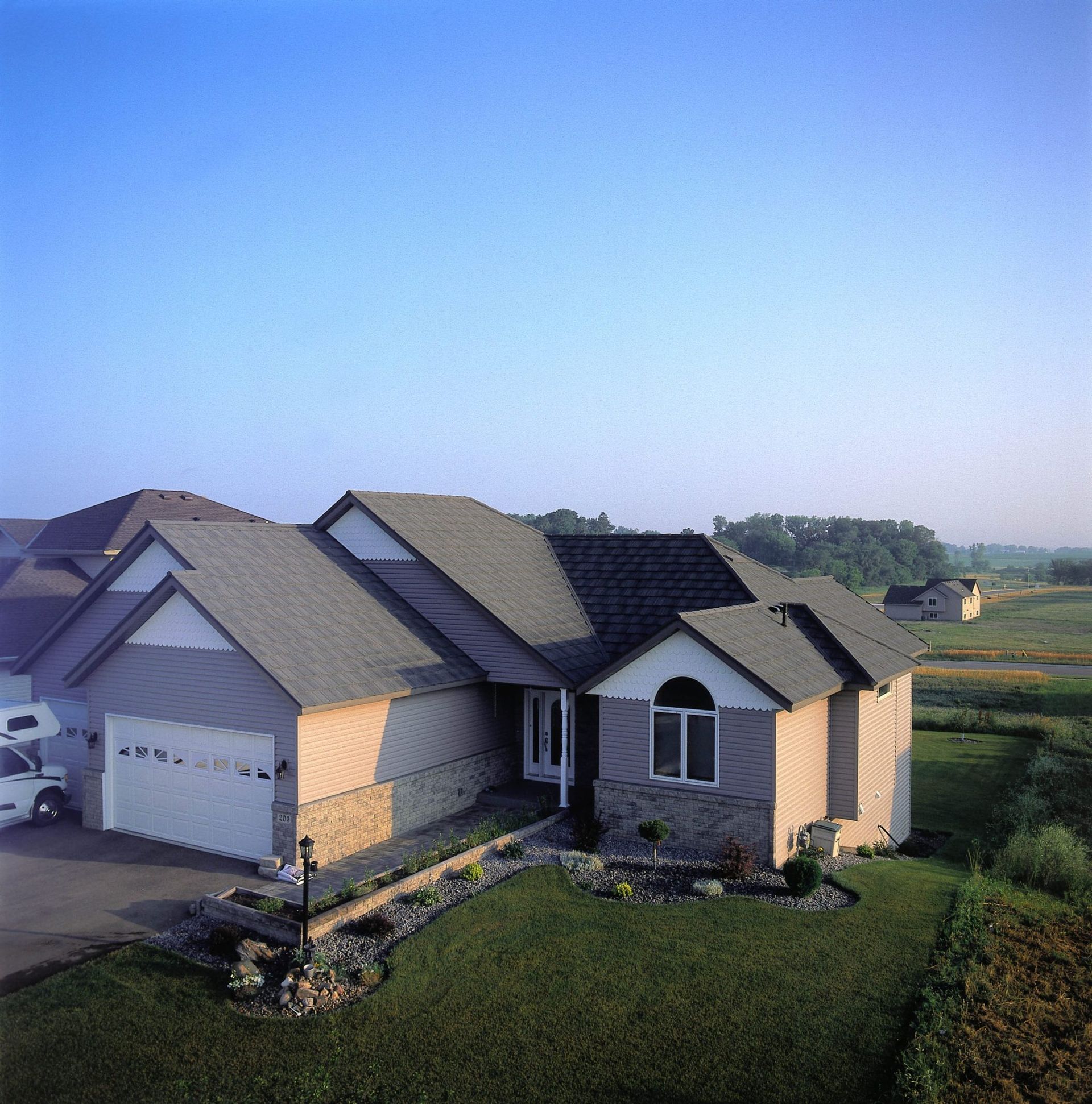 A house with a garage, light brick siding, and a dark roof sits on a grassy hill under a blue sky.