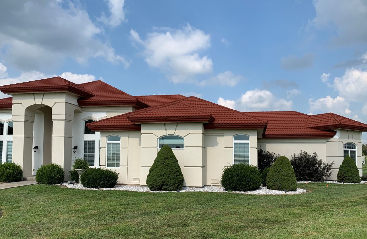 Large cream-colored house with a red tile roof against a blue sky. Well-manicured lawn and bushes in front.