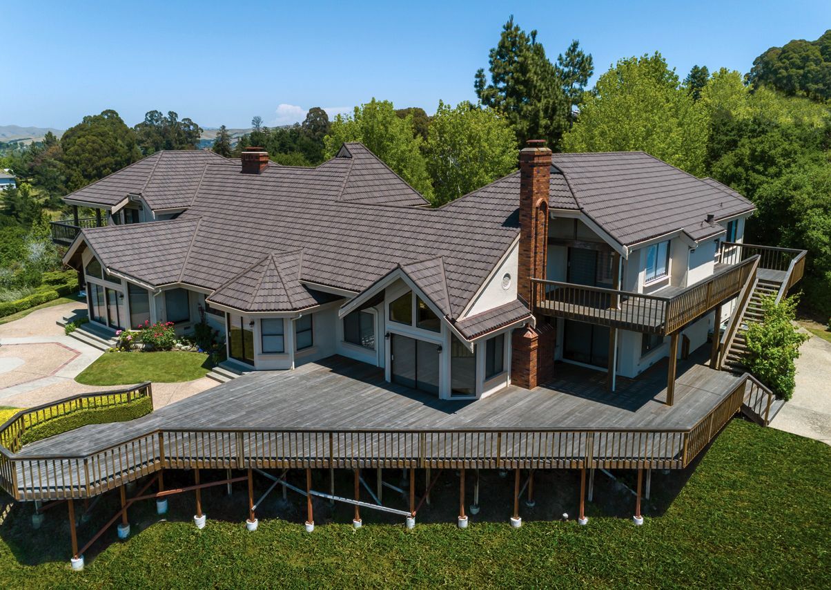 Aerial view of a large house with a dark roof and large wooden deck surrounded by green grass and trees.
