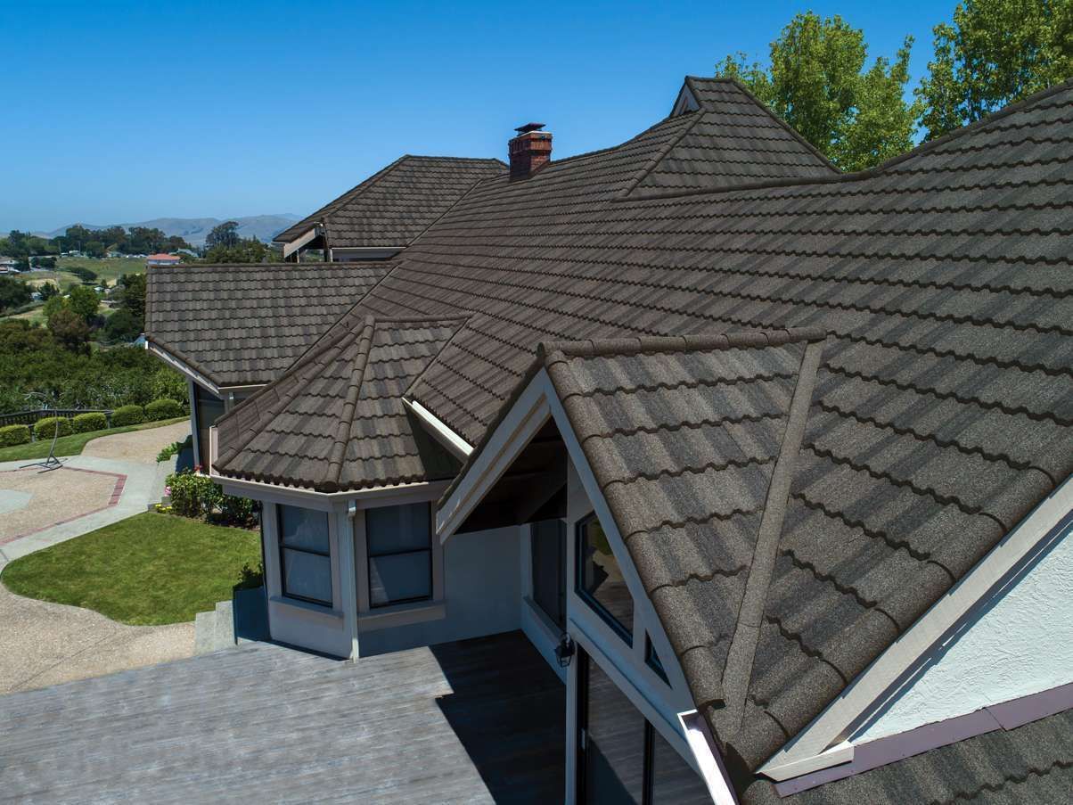 An aerial view of a house with a dark brown shingle roof, under a bright blue sky.