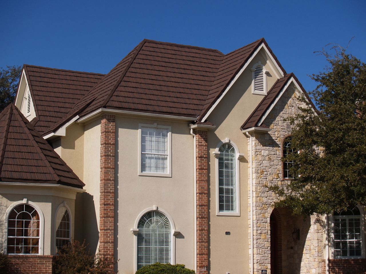 Two-story house with brown roof, beige walls, brick and stone accents, and white-framed windows against a blue sky.
