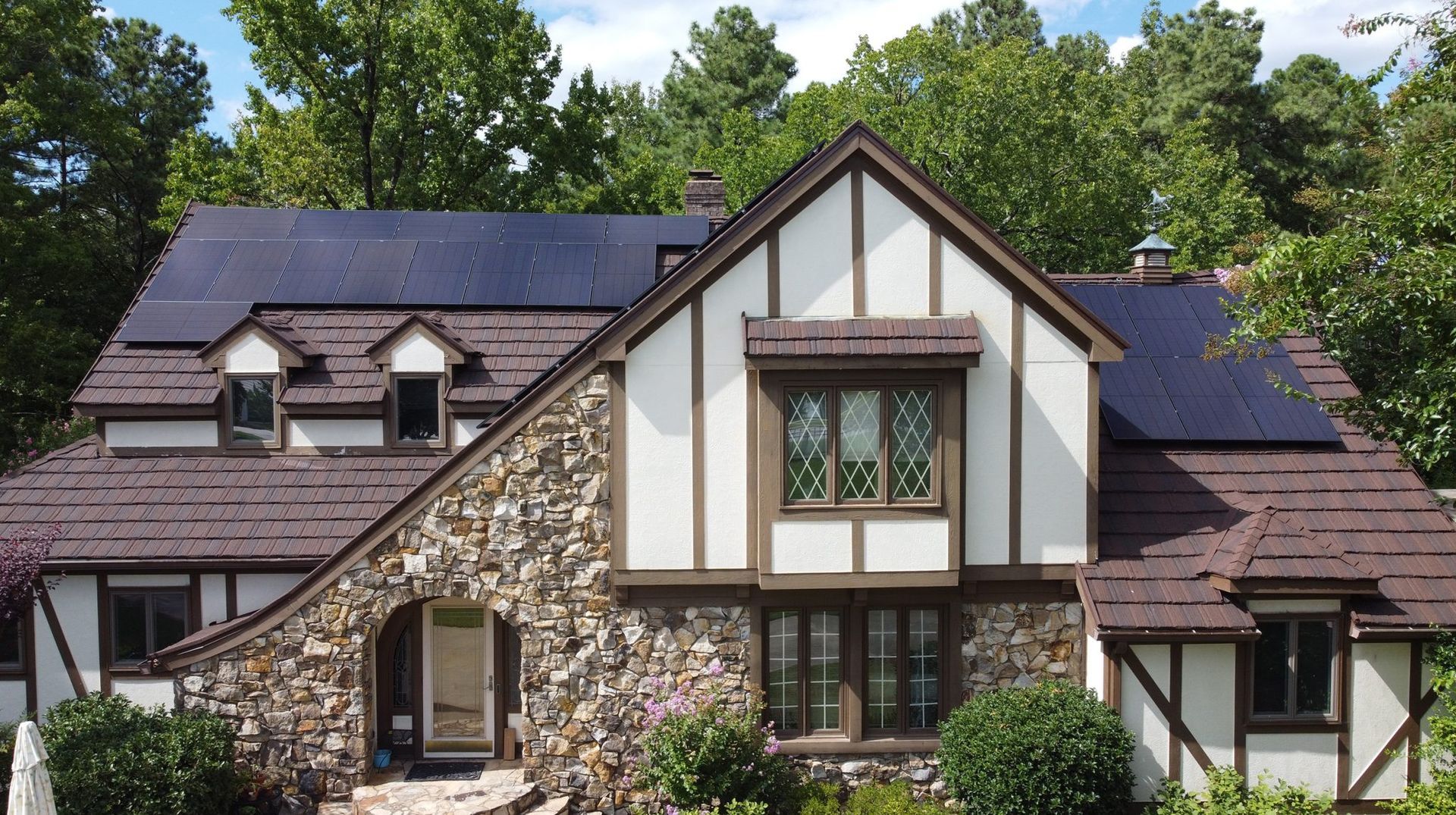 Tudor-style house with solar panels on the roof, brown and white exterior, surrounded by green trees under a blue sky.
