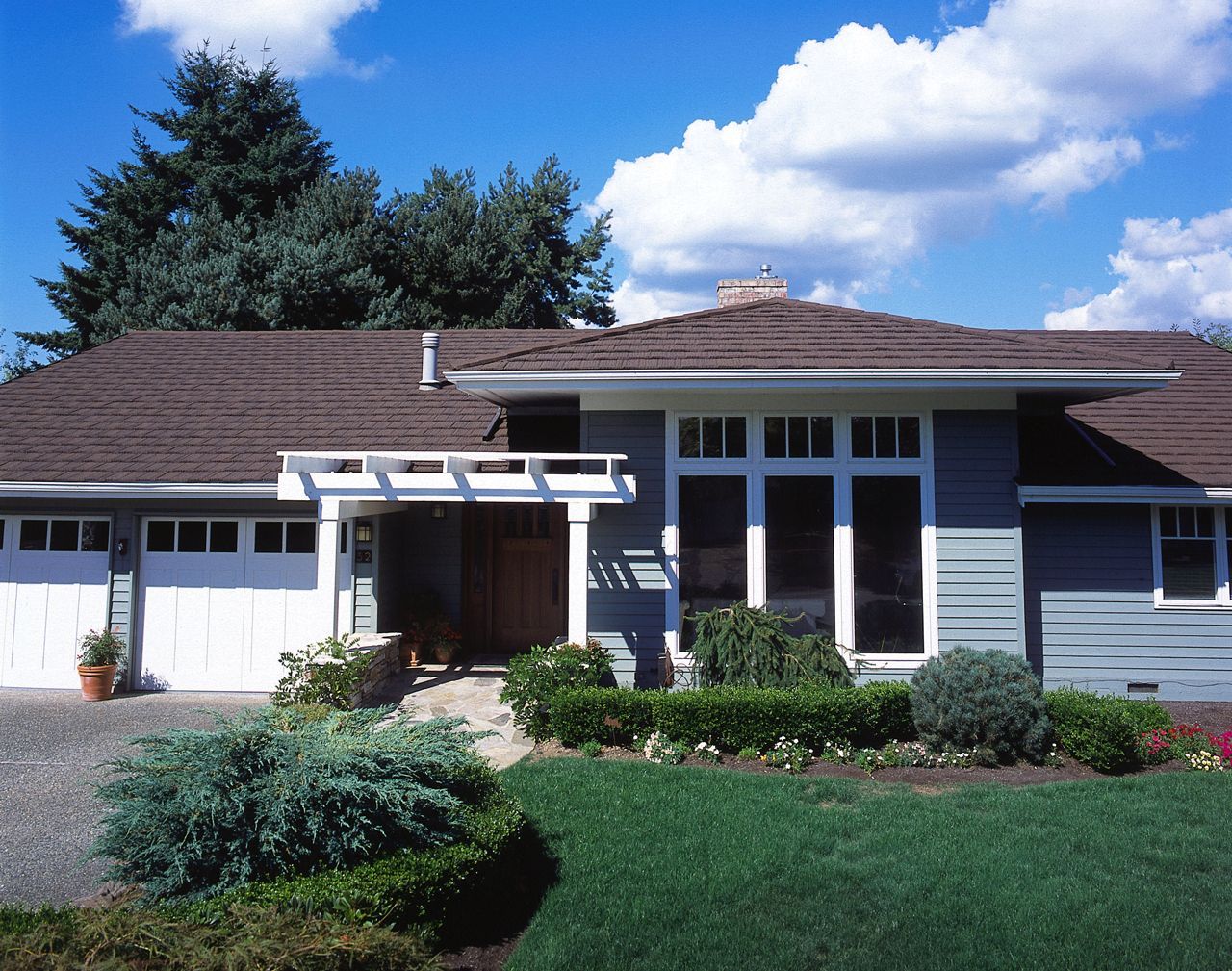 A blue house with a brown roof, white trim, and a well-kept yard. A white garage door and front entryway are visible.