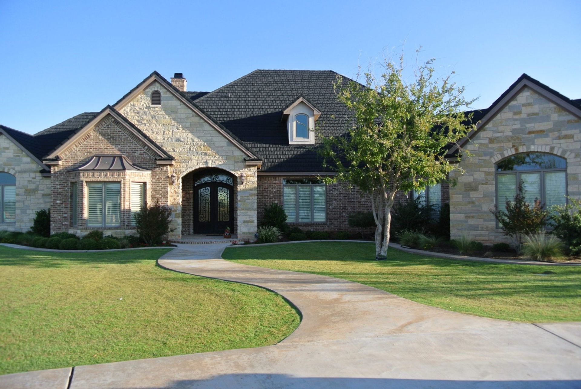 House with stone and brick facade, surrounded by green lawn, with a curved driveway leading to the front door.