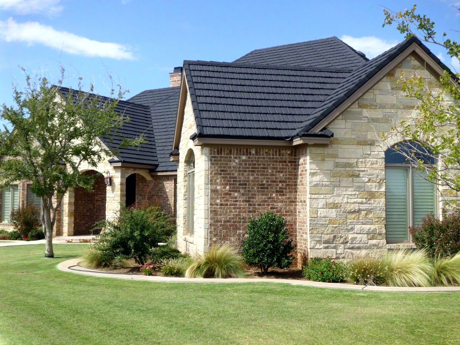 House with stone and brick exterior, dark gray roof, and well-manicured lawn under a blue sky.