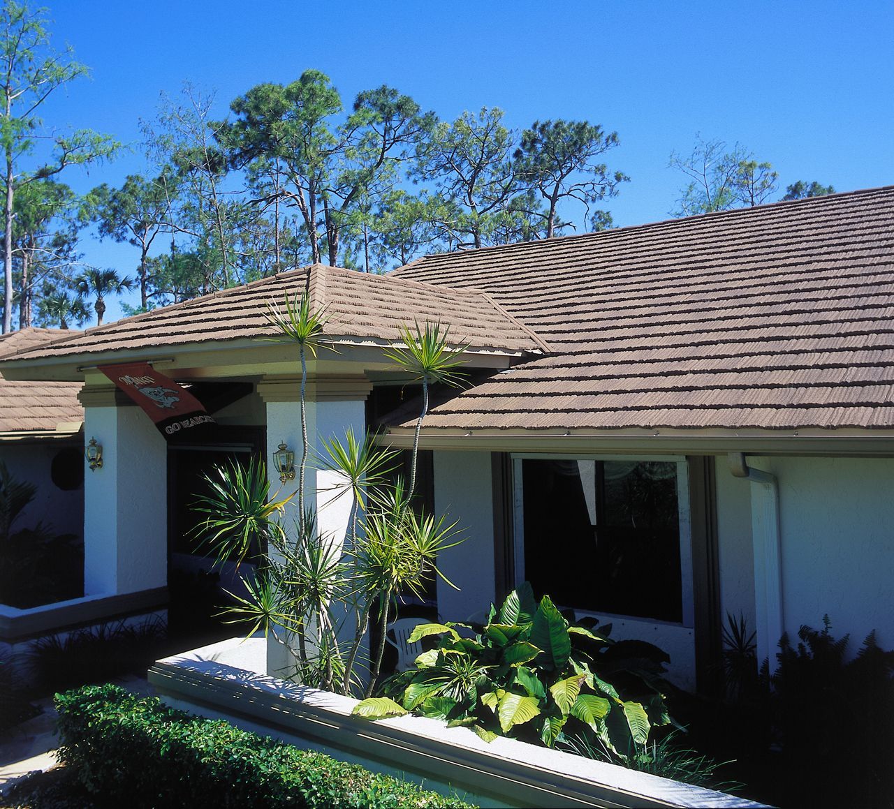 A beige house with a brown tiled roof under a bright blue sky. Plants and bushes in front of the building.
