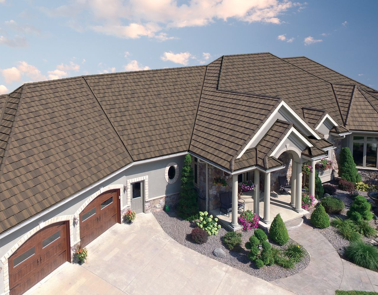 A large, multi-gabled house with brown shingled roof, attached garage with brown doors, and well-manicured landscaping on a sunny day.