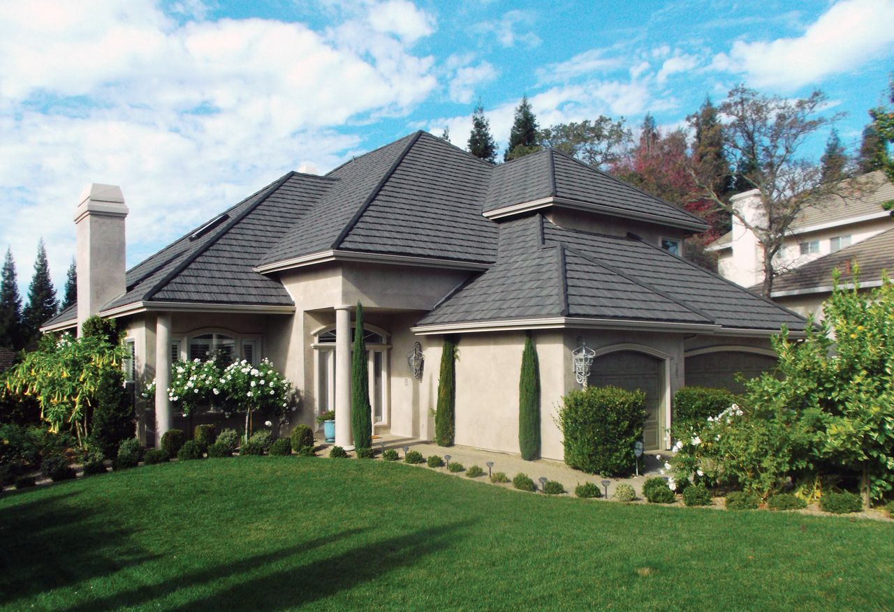 A beige house with a dark gray roof, lush green lawn, and tall trees under a blue sky with clouds.