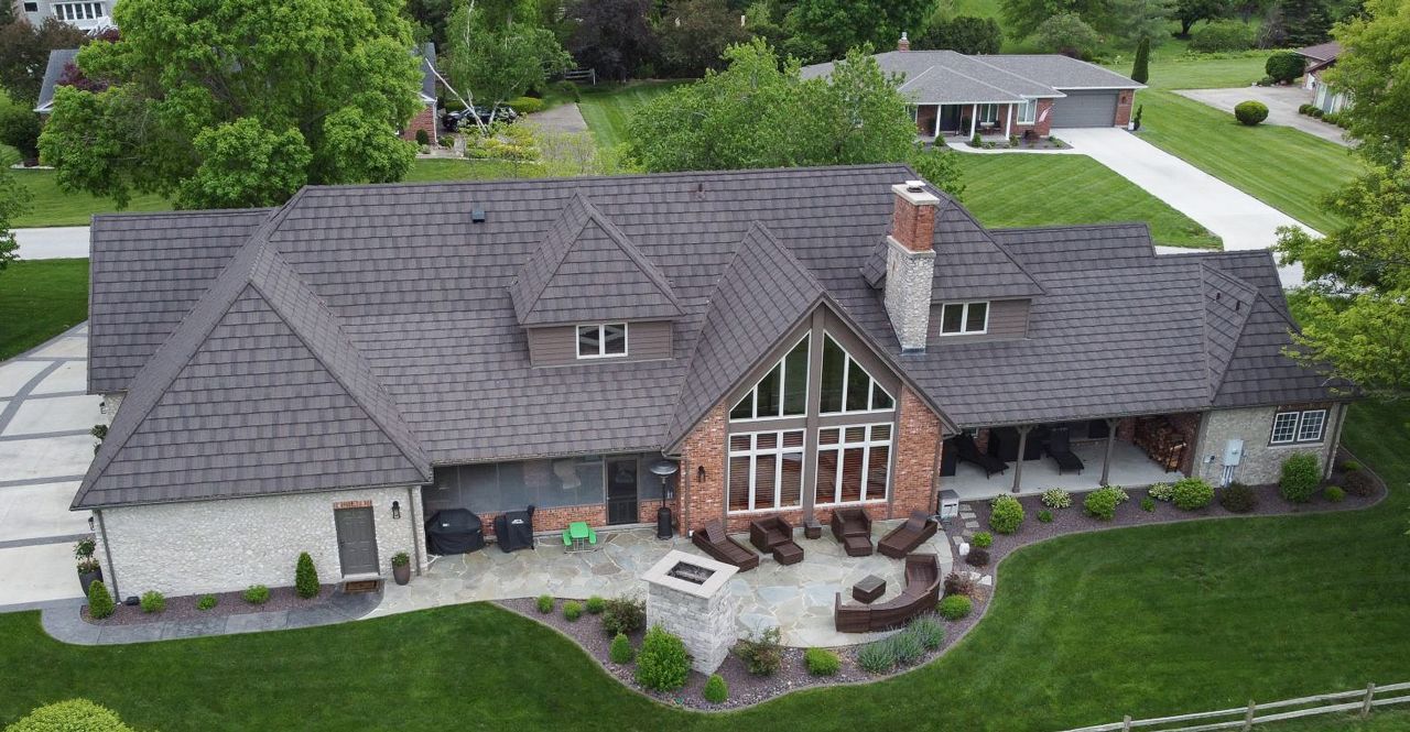 An aerial view of a large brick house with a dark gray shingle roof, a patio, and a green lawn.