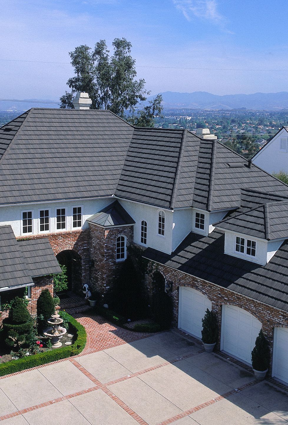 Large white house with a gray tile roof, brick accents, and a concrete driveway. Greenery and a city view are in the background.