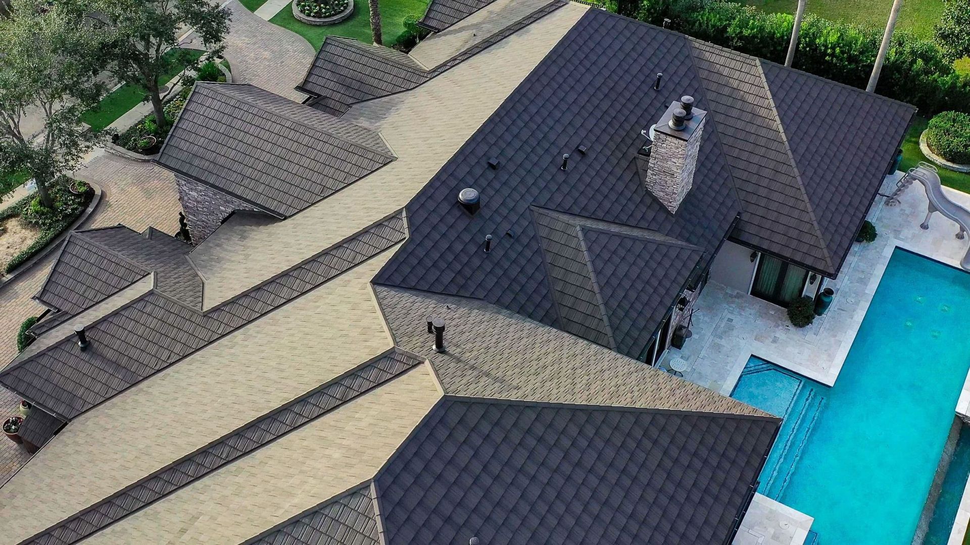 Aerial view of a multi-level house with a swimming pool. The roof is dark gray, and the house is surrounded by greenery.