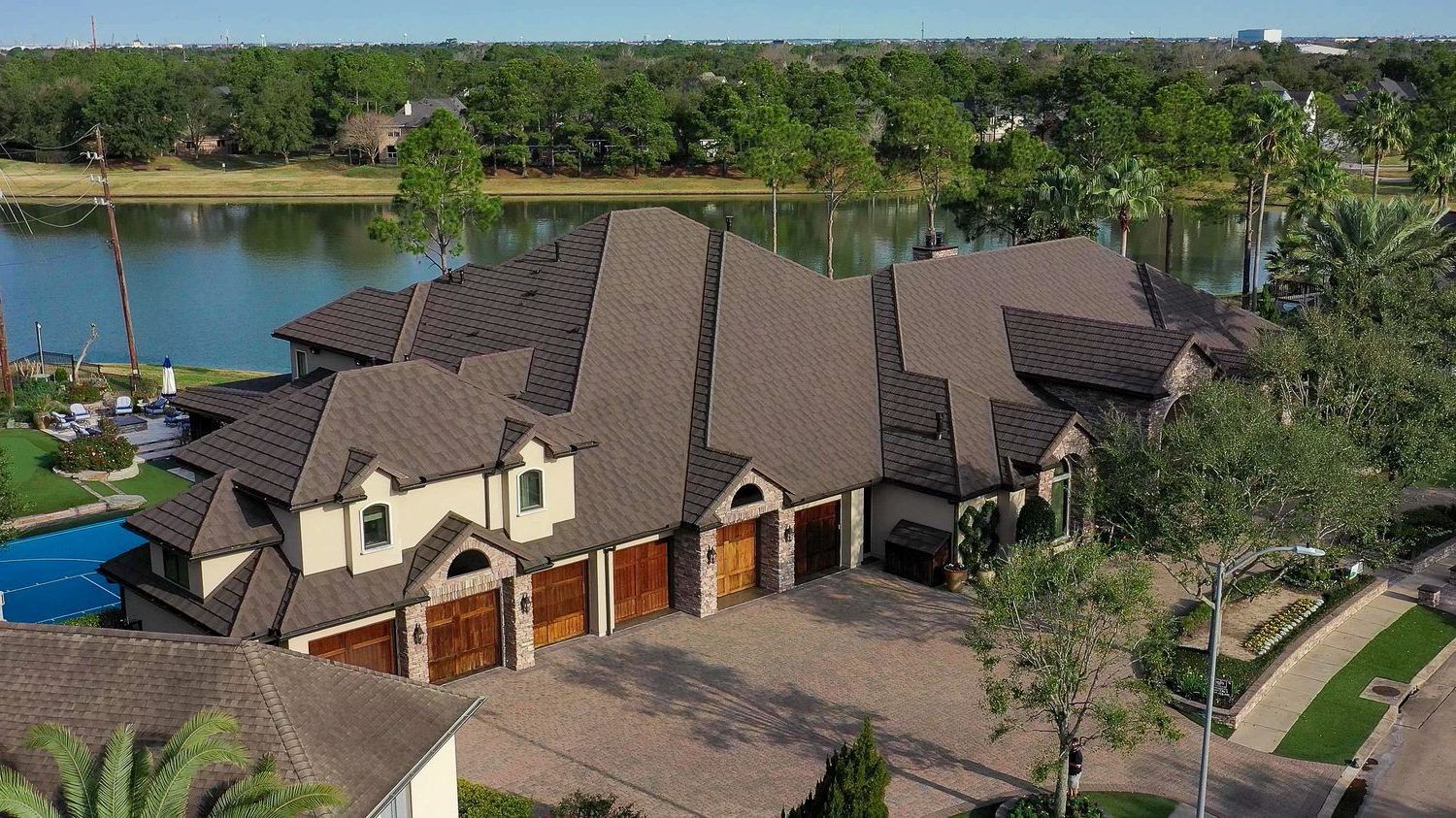 Large luxury home on a lake with a driveway and multiple garage doors. Brown roof, beige exterior, and trees surround the property.