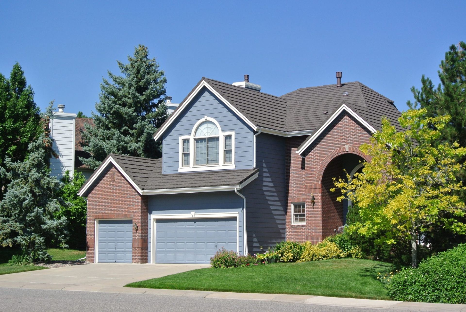 Two-story blue house with red brick accents, two-car garage, and arched entryway, on a sunny day.