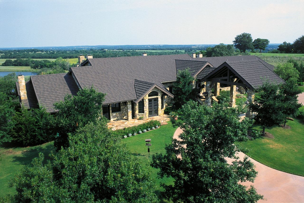 Large, stone-clad home with brown roof, surrounded by trees and a circular driveway, overlooking a distant landscape.