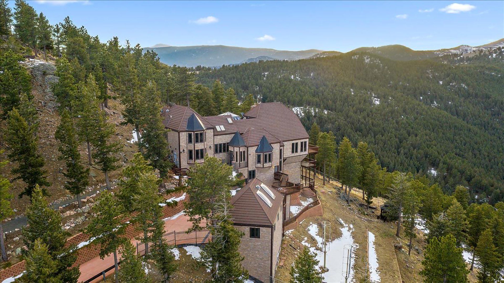Large house on a hillside with a forest, mountain views, and patches of snow.