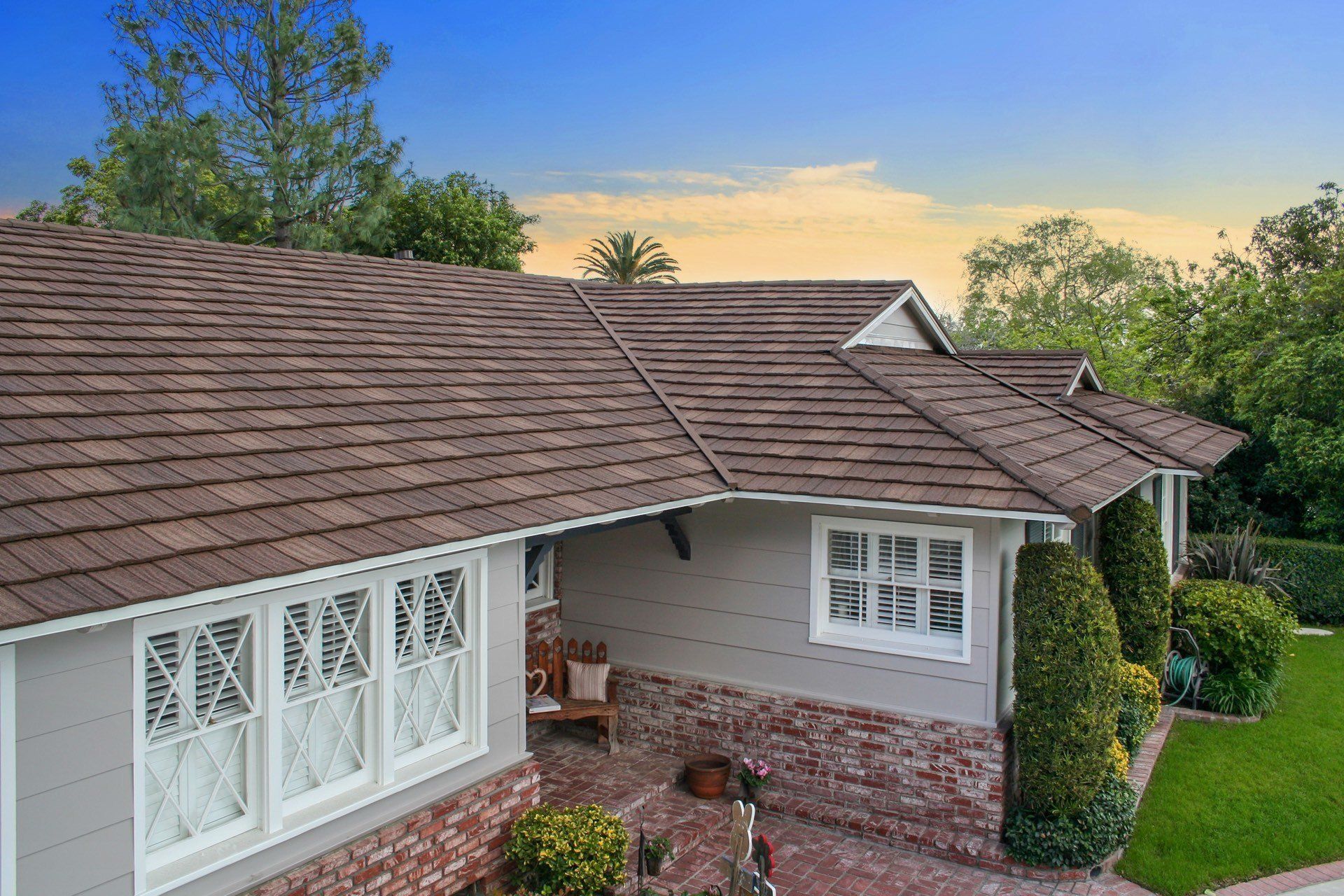 A brown-shingled roof covers a light gray house with white-trimmed windows and brickwork, set against a blue sky and green trees.