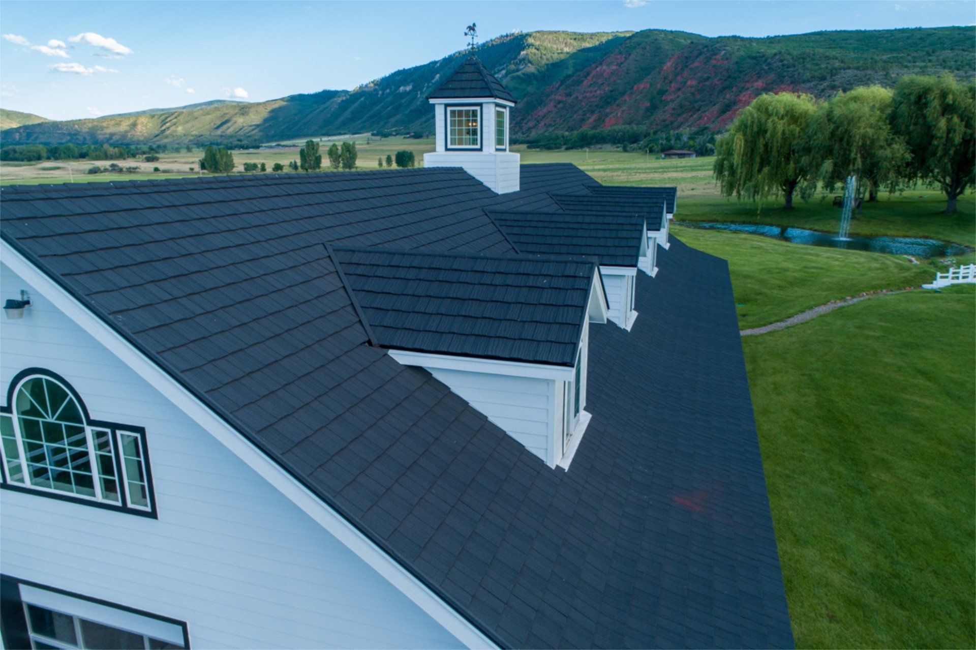 White barn with a dark blue roof and dormers, set against a backdrop of mountains and a green meadow.