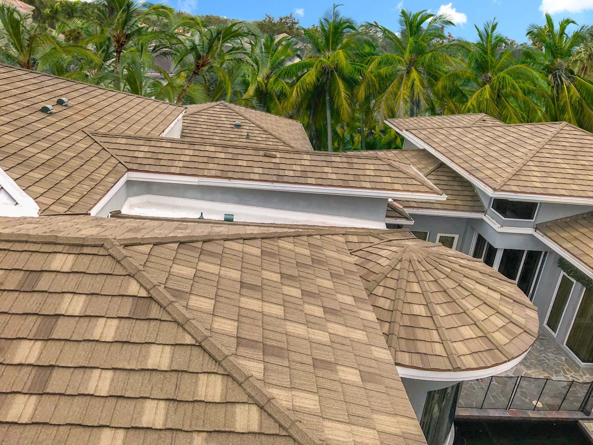 Tan shingled rooftops of a multi-level house against a backdrop of green palm trees and blue sky.