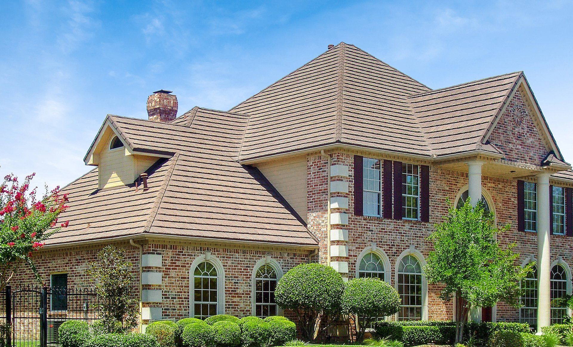Two-story house with a brown roof, brick and beige exterior, arched windows, and manicured lawn under a blue sky.