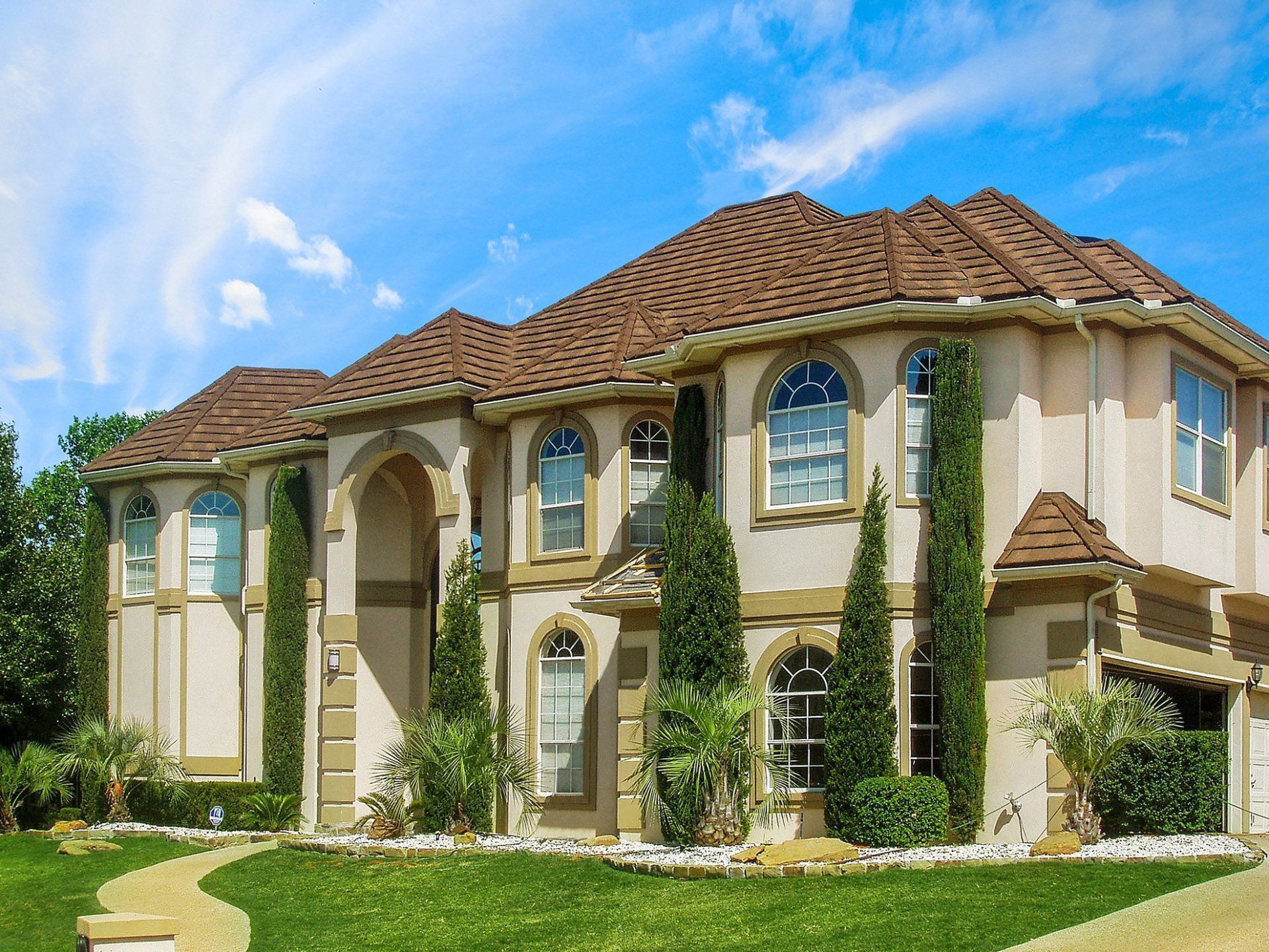 Large, two-story beige house with arched windows and a brown tile roof, set against a blue sky with a manicured lawn.
