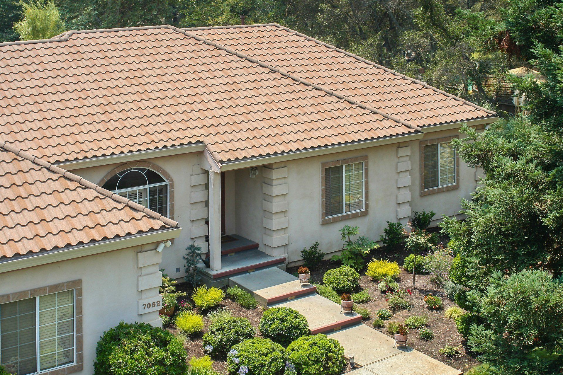 Tan-roofed house with a tan exterior and a walkway leading to the front door, surrounded by landscaping and trees.