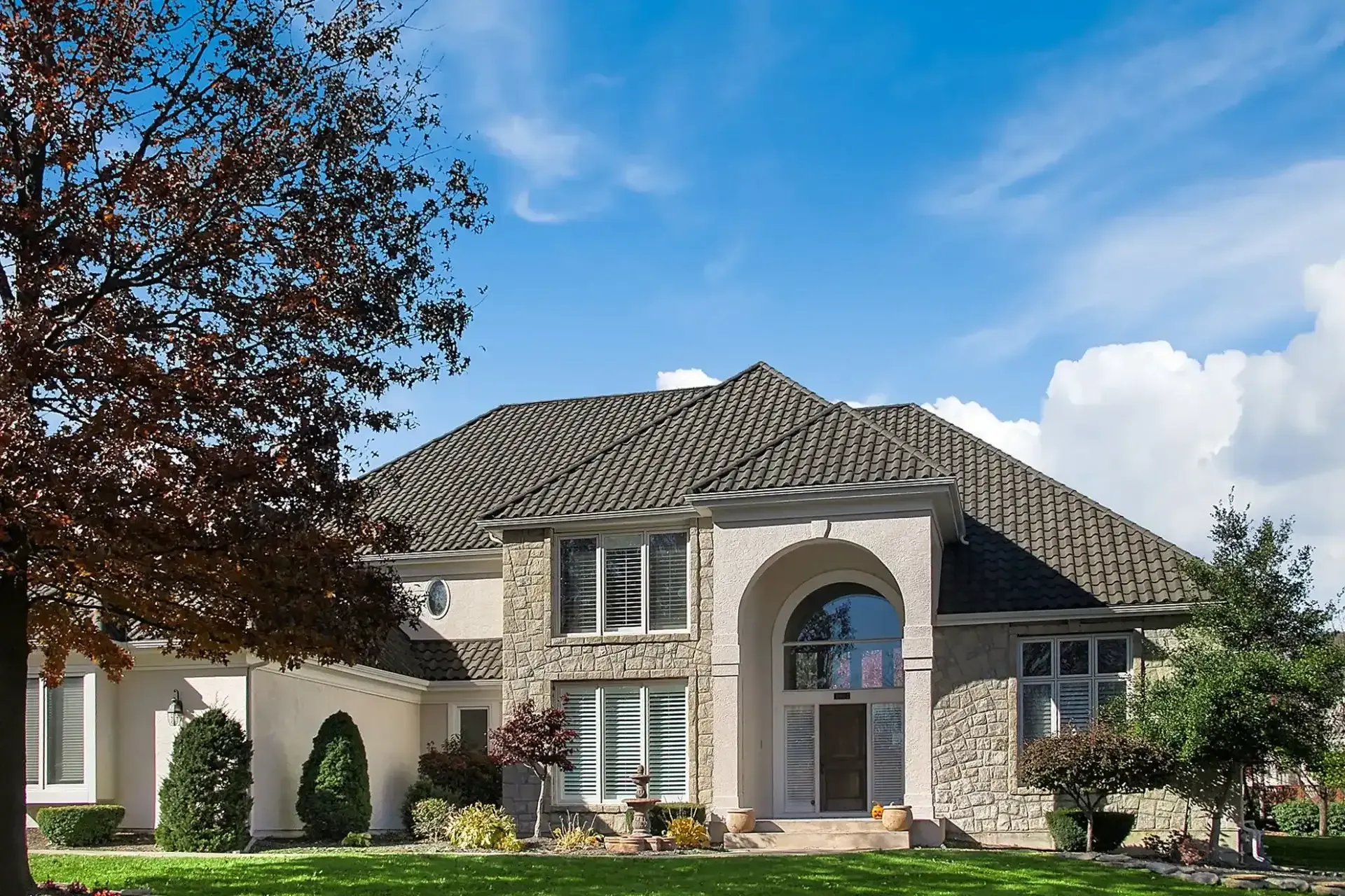 Two-story beige and stone house with a tiled roof, set against a blue sky with clouds, framed by trees and green grass.