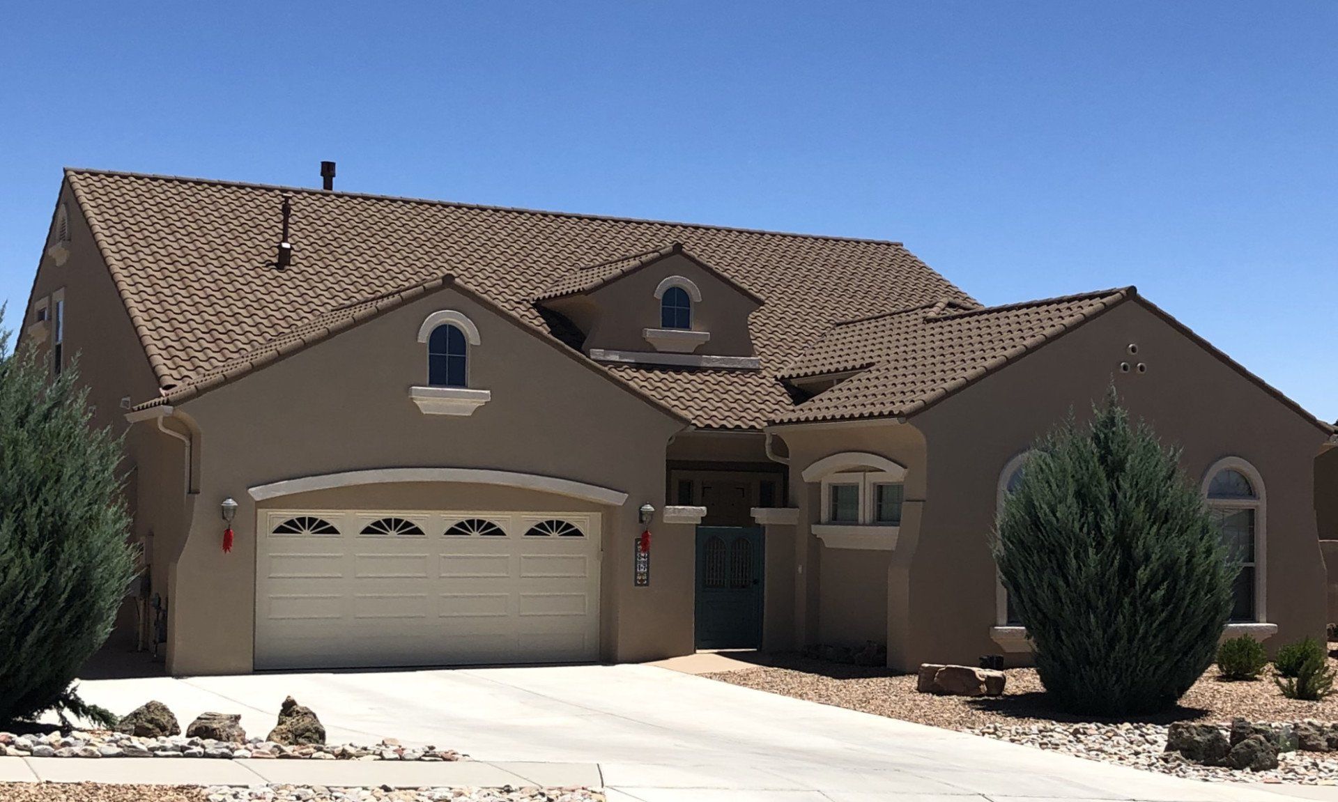 Tan stucco house with beige garage door and brown tile roof under a clear blue sky. Green bushes flank the driveway.