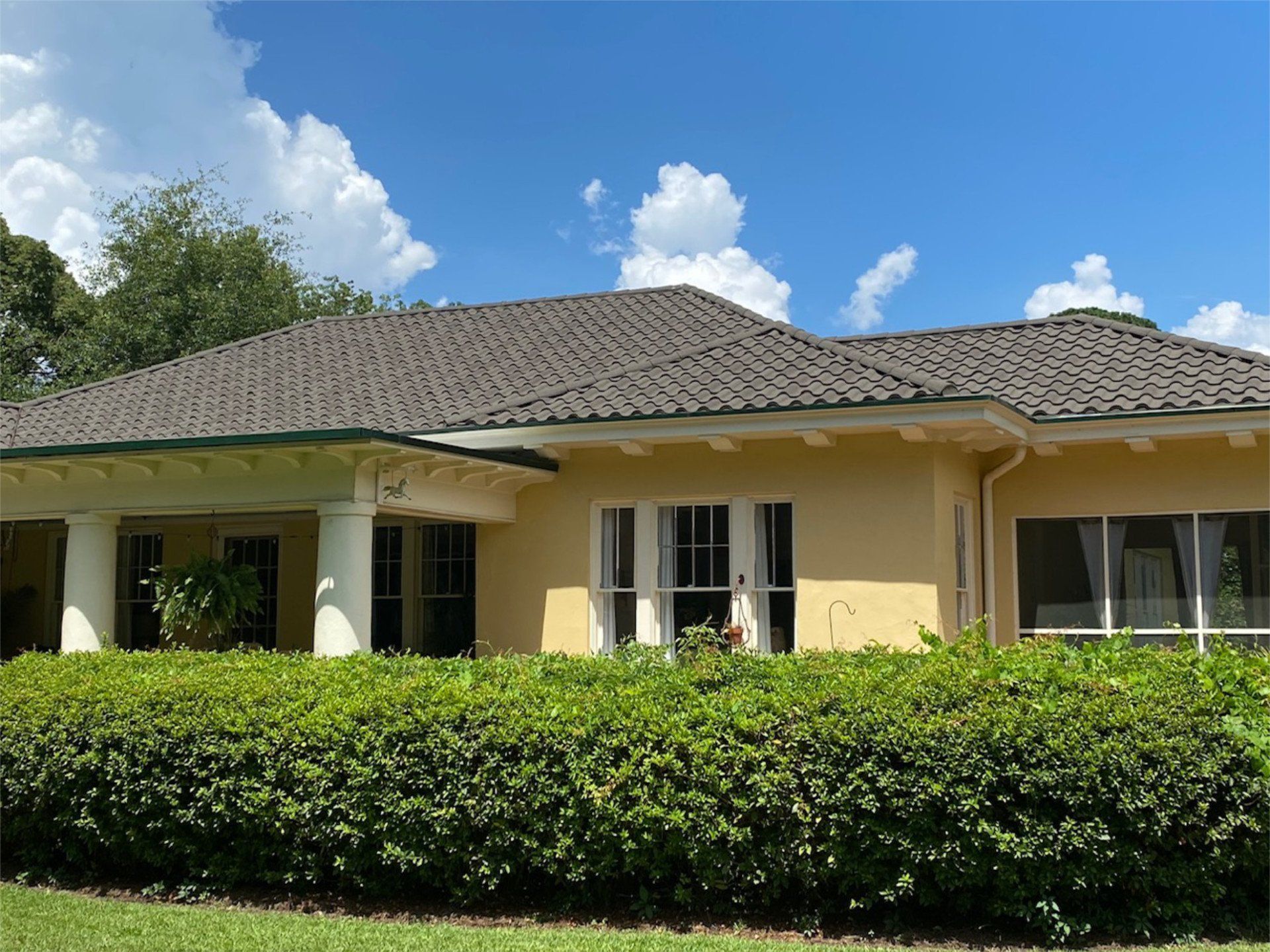 Yellow stucco house with dark roof and white columns, fronted by a green hedge under a partly cloudy blue sky.