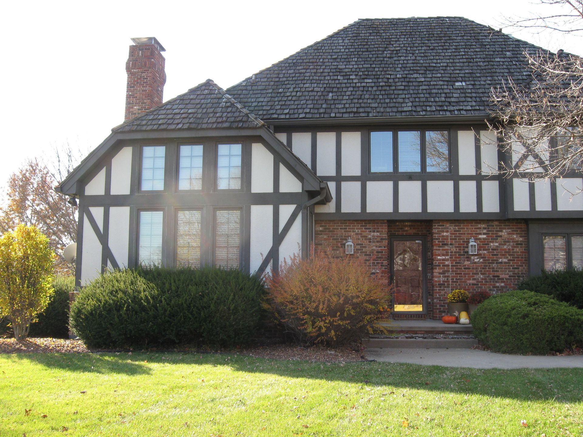 Tudor-style house with dark trim, brick, and a textured roof. Green bushes and grass in front.