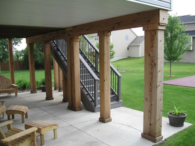 Covered patio with wooden support columns, stairs, and furniture. The setting is a backyard with a lawn.