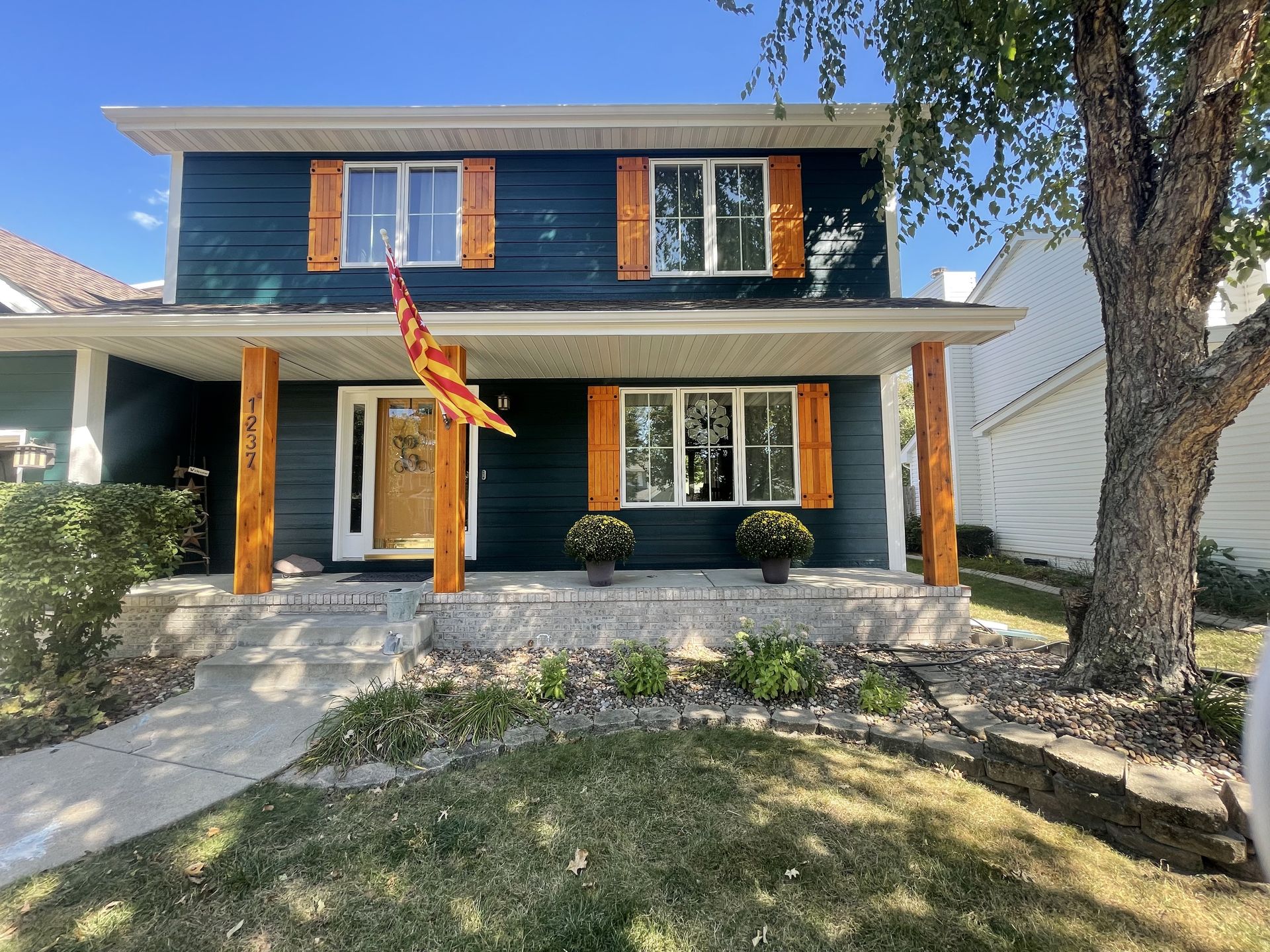 Two-story blue house with wood shutters and pillars. A flag hangs near the front door, and fall flower pots sit on the porch.